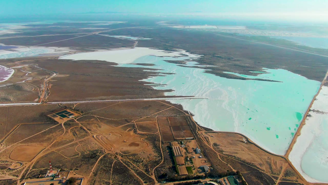 High aerial frame over Guerrero Negro, Baja California Sur, with turquoise and white salt evaporation ponds on the right, brown arid terrain and roads on the left, and no visible people.