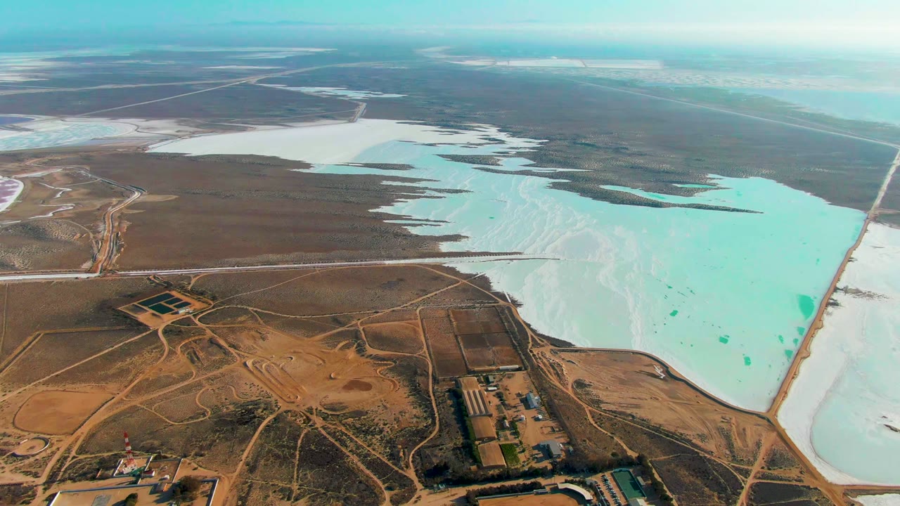 Drone-height frame over Guerrero Negro with pale blue saline water, white salt deposits, a long straight road or levee at the pond edge, dry brown flats, and no visible people.
