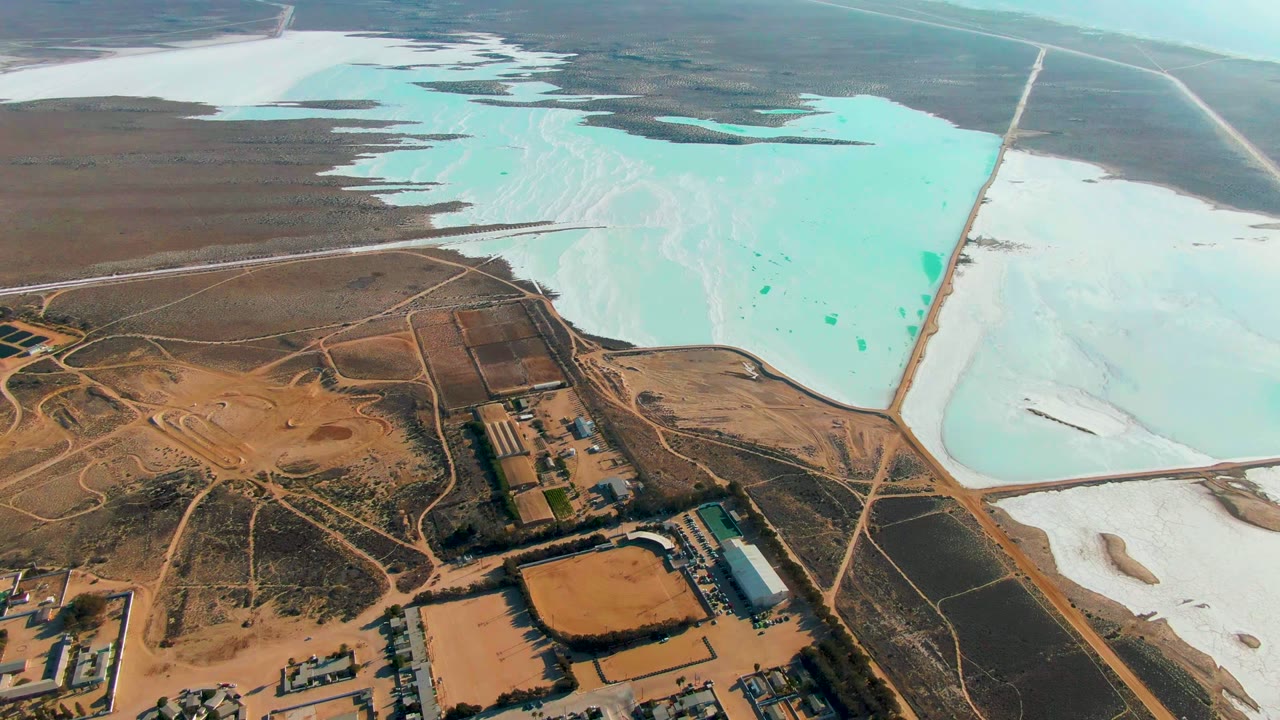 Opening aerial frame over Guerrero Negro, Baja California Sur, with turquoise evaporation ponds, white salt areas, brown desert ground, facility buildings, and no visible people.