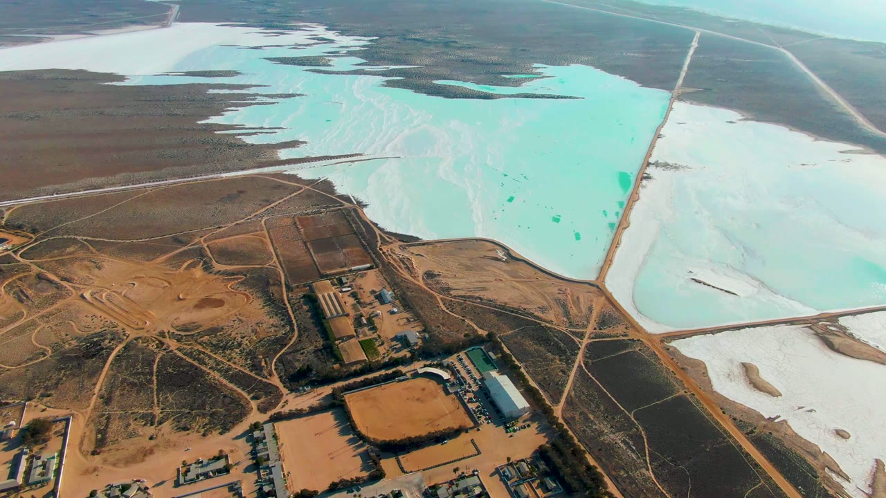 Mid-clip aerial frame over Guerrero Negro salt ponds, with facility buildings near the lower edge, pale salt flats to the right, turquoise pond water, and no visible people.