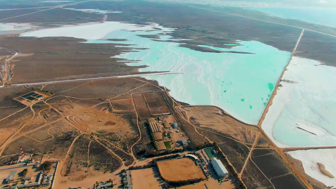 Opening aerial frame over Guerrero Negro, Baja California Sur, with turquoise salt evaporation ponds beyond brown industrial land, dirt roads, buildings, and no visible people.