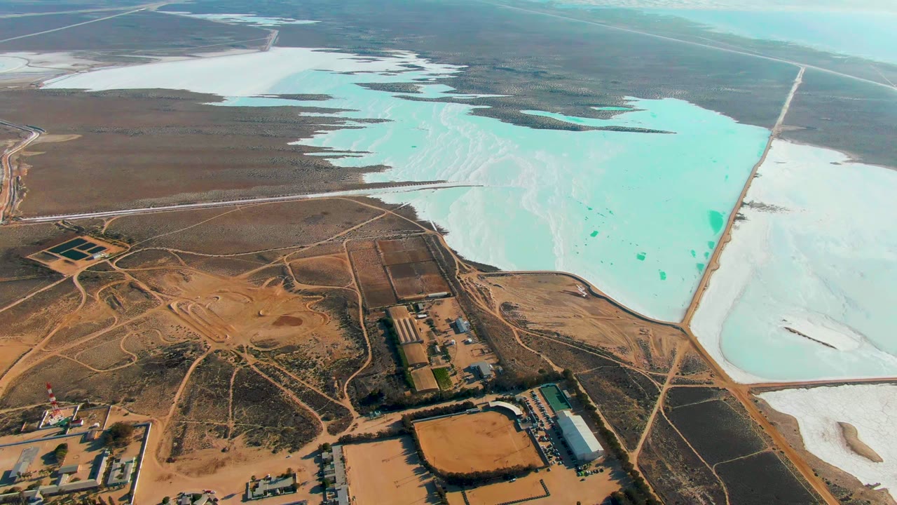 High frame over Guerrero Negro salt ponds with a large turquoise basin at center right, white mineral deposits to the side, industrial lots below, and no visible people.