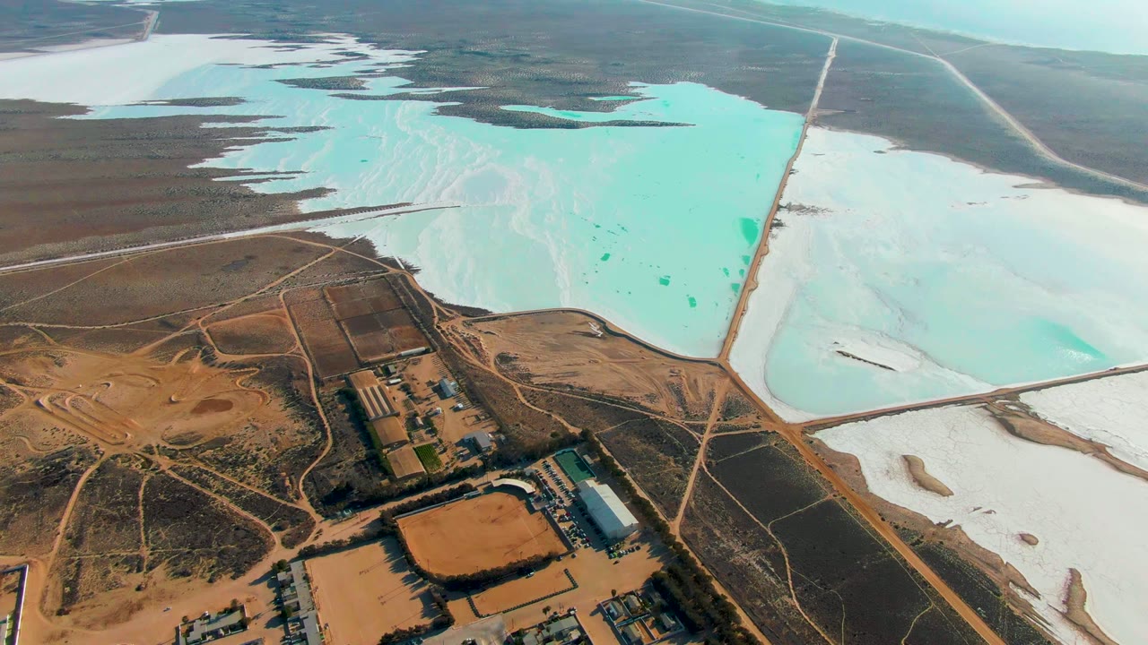 Opening aerial frame over Guerrero Negro evaporation ponds, with turquoise water and white salt flats above brown roads and industrial buildings, no visible people.
