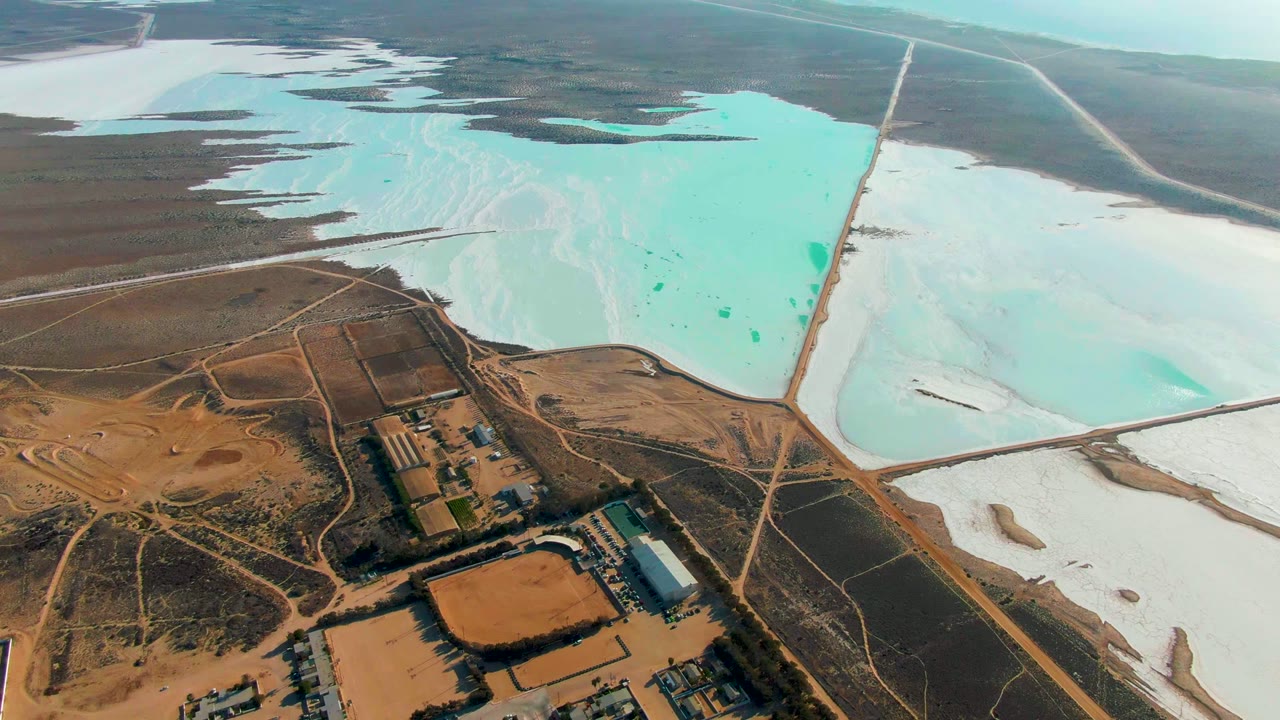 Drone view over pale salt ponds and brown desert roads beside an industrial compound.