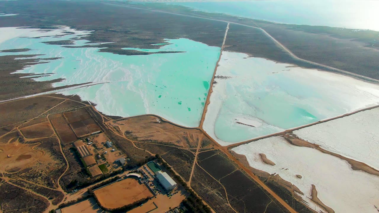 Wide aerial view over salt evaporation ponds in Guerrero Negro, Baja California Sur, with turquoise water on the left, white salt flats on the right, dry terrain below, and no visible people.