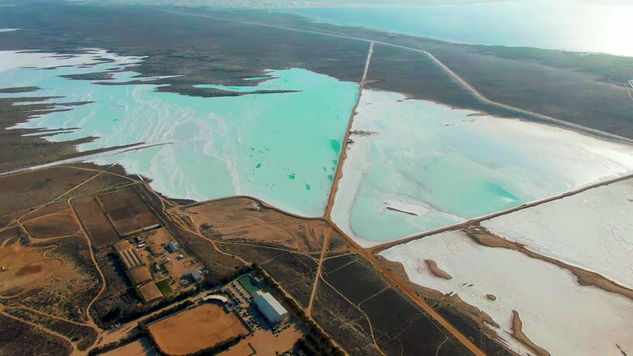 Aerial salt ponds with turquoise water, white salt flats, and industrial buildings below.
