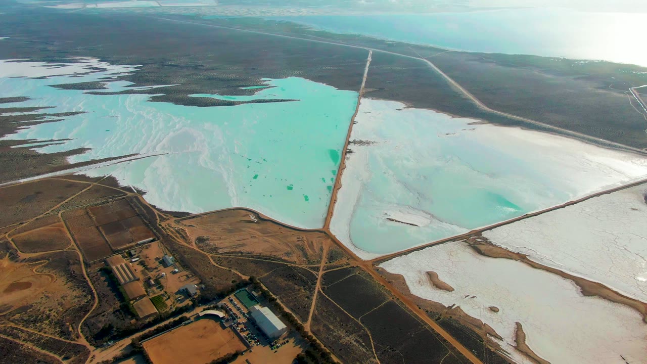 Oblique aerial view in Guerrero Negro with turquoise evaporation water across the left pond, pale white salt on the right, crossing roads through the center, distant flat land, and no visible people.