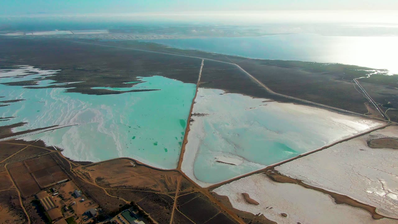 High aerial frame over Guerrero Negro salt evaporation ponds with turquoise water at left, white halite flats at right, distant water on the horizon, and no visible people.