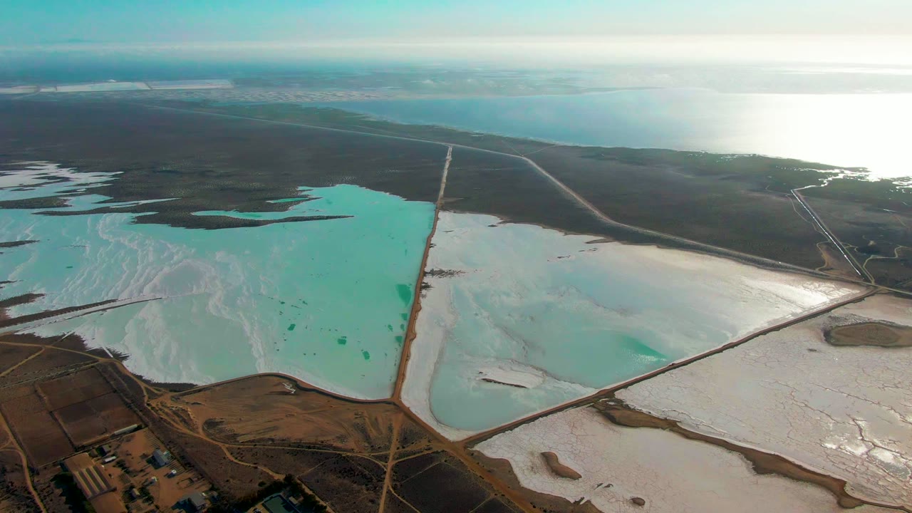 Drone view of geometric salt evaporation ponds with turquoise water and pale white deposits.