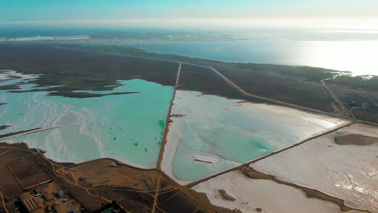 Wide drone frame over Guerrero Negro evaporation ponds with a turquoise basin crossing the lower left, white halite on the lower right, brown levees, distant water, and no visible people.