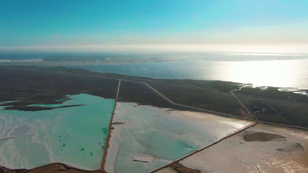 Aerial frame over Guerrero Negro salt ponds with turquoise basins and pale white deposits in the foreground, dark terrain beyond, bright water on the horizon, and no visible people.