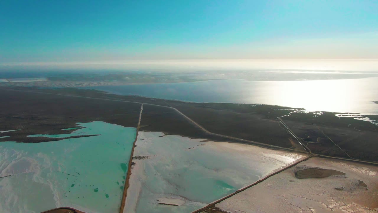Wide aerial view of salt ponds divided by levees with bright reflection on distant water.