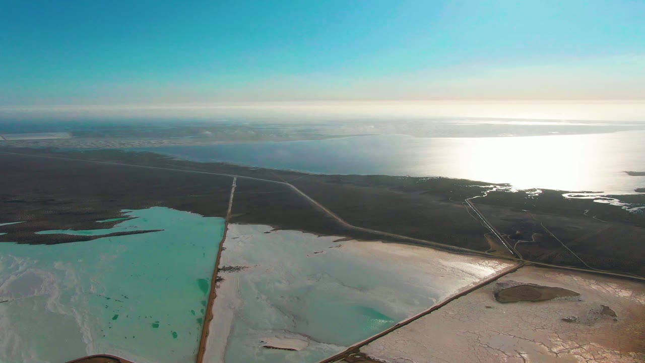 Guerrero Negro aerial frame with turquoise salt pond water on the left, pale salt deposits below, dark terrain and road lines in the midground, bright distant water, and no visible people.