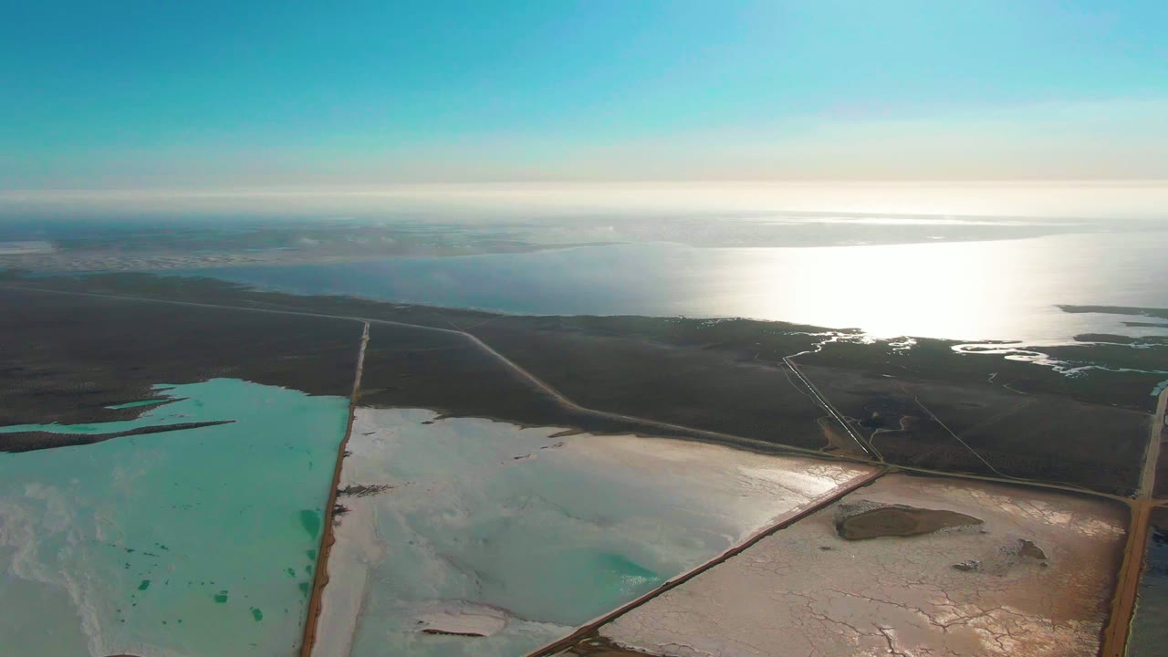 Aerial view over Guerrero Negro salt evaporation ponds, with turquoise water at left, pale salt flats below, straight roads crossing the ponds, bright ocean glare beyond, and no visible people.