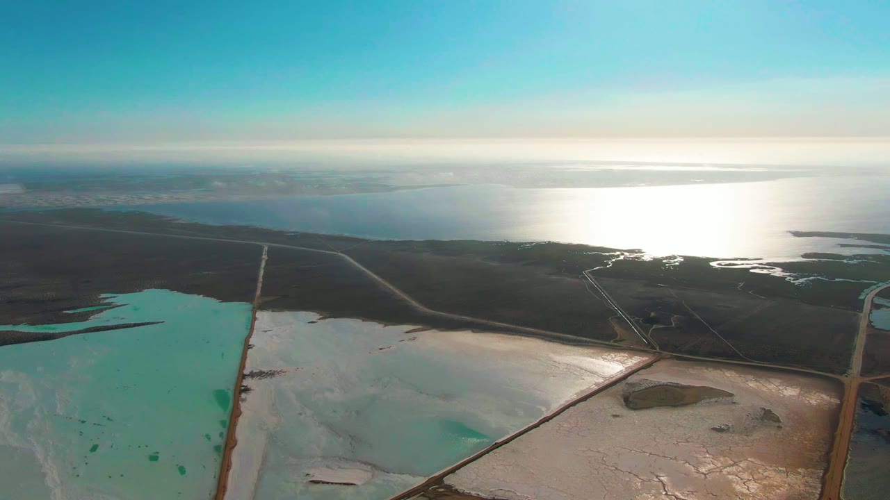 Wide aerial of salt ponds, service roads, dark land, and bright ocean horizon.