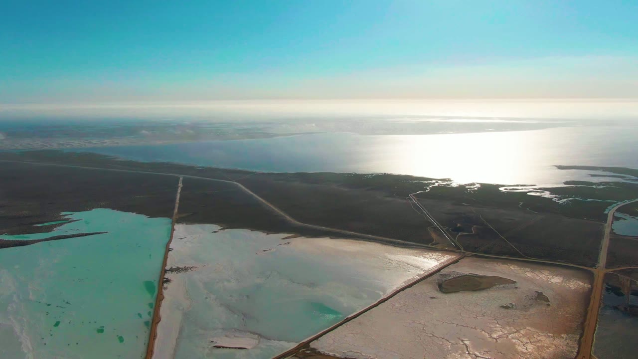 Guerrero Negro evaporation ponds fill the lower frame with turquoise and pale water, while road lines meet near the center and bright reflected sunlight spreads across the ocean in the background, with no visible people.