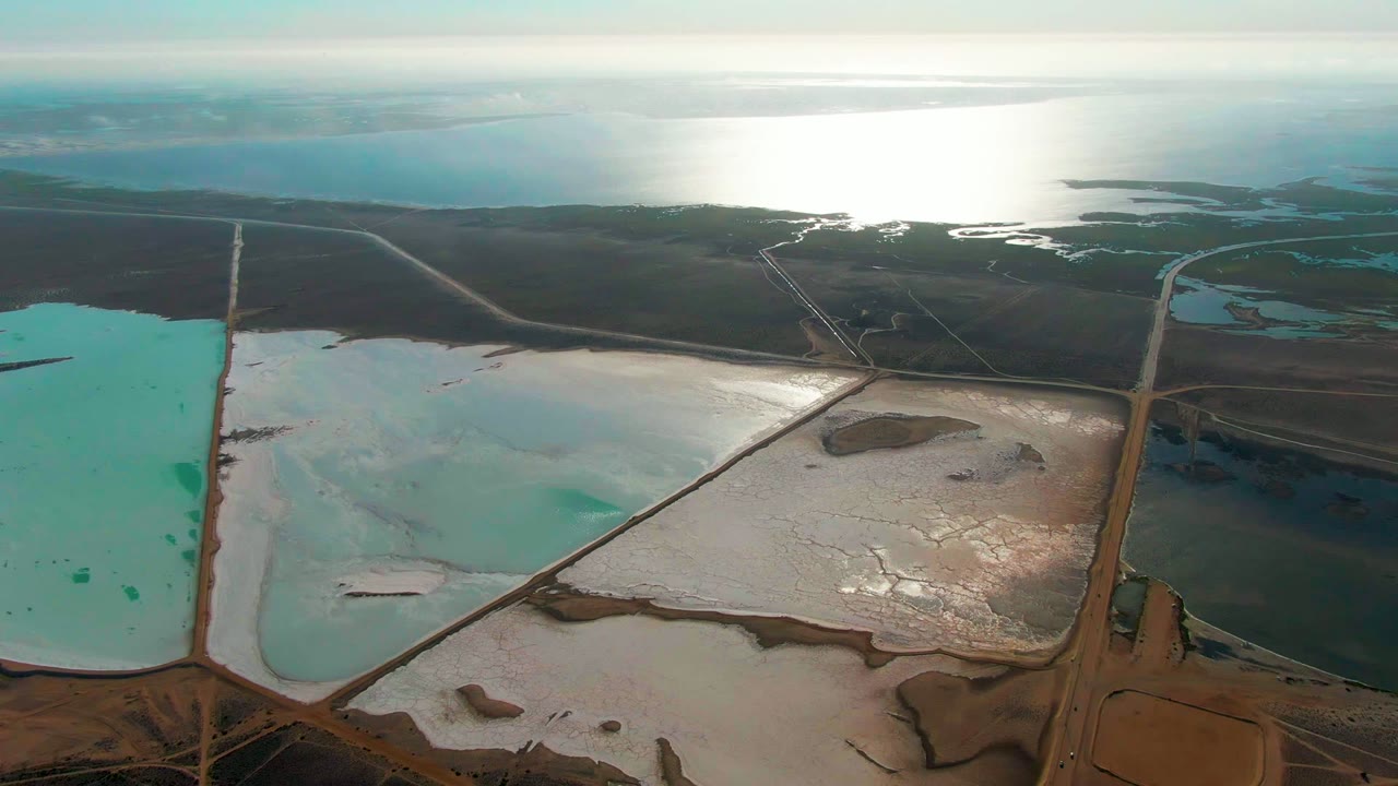 Aerial view over Guerrero Negro salt evaporation ponds with a turquoise pool at left, white salt flats in the center, brown roads crossing the foreground, distant reflective water, and no visible people.