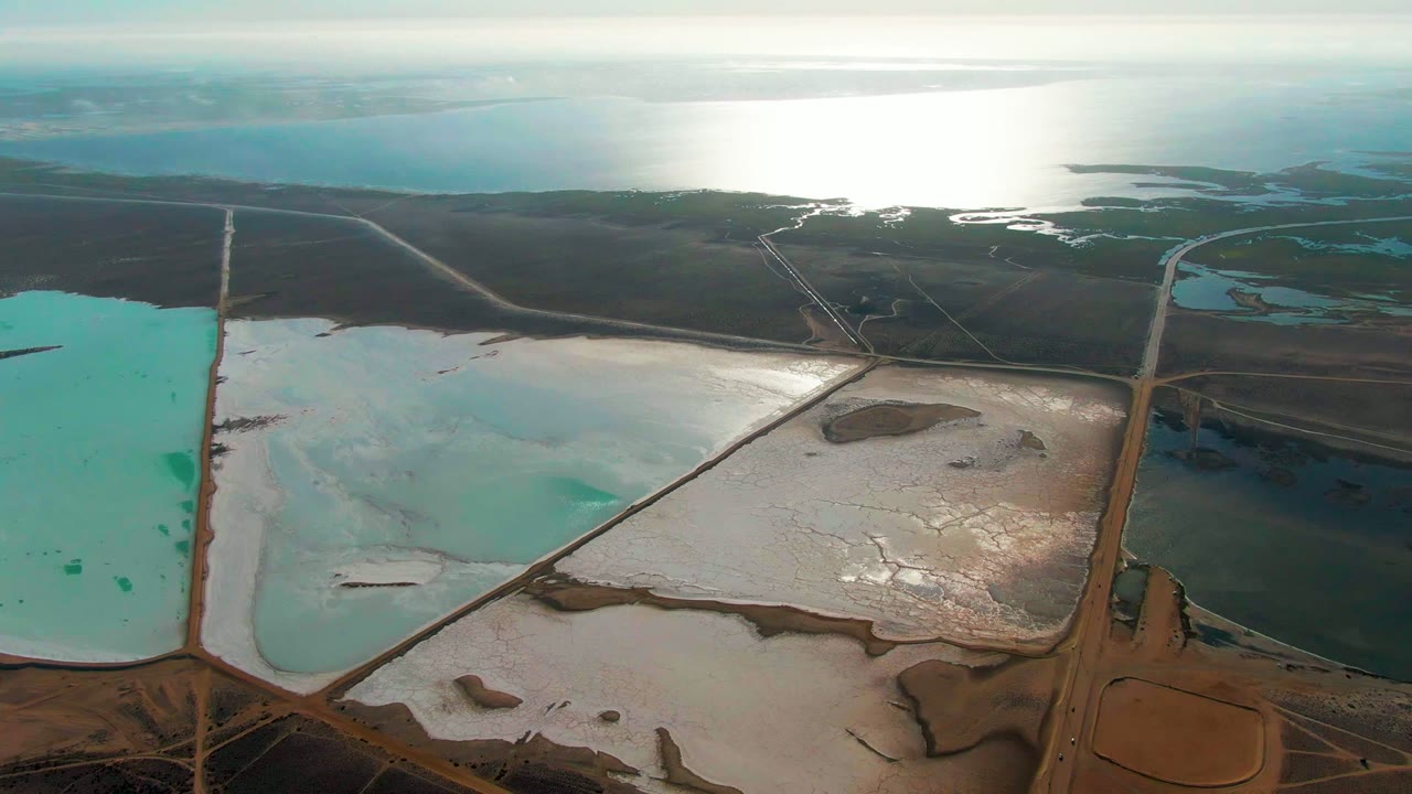 Aerial salt ponds with turquoise water, white deposits, diagonal levees, and coastal water beyond.