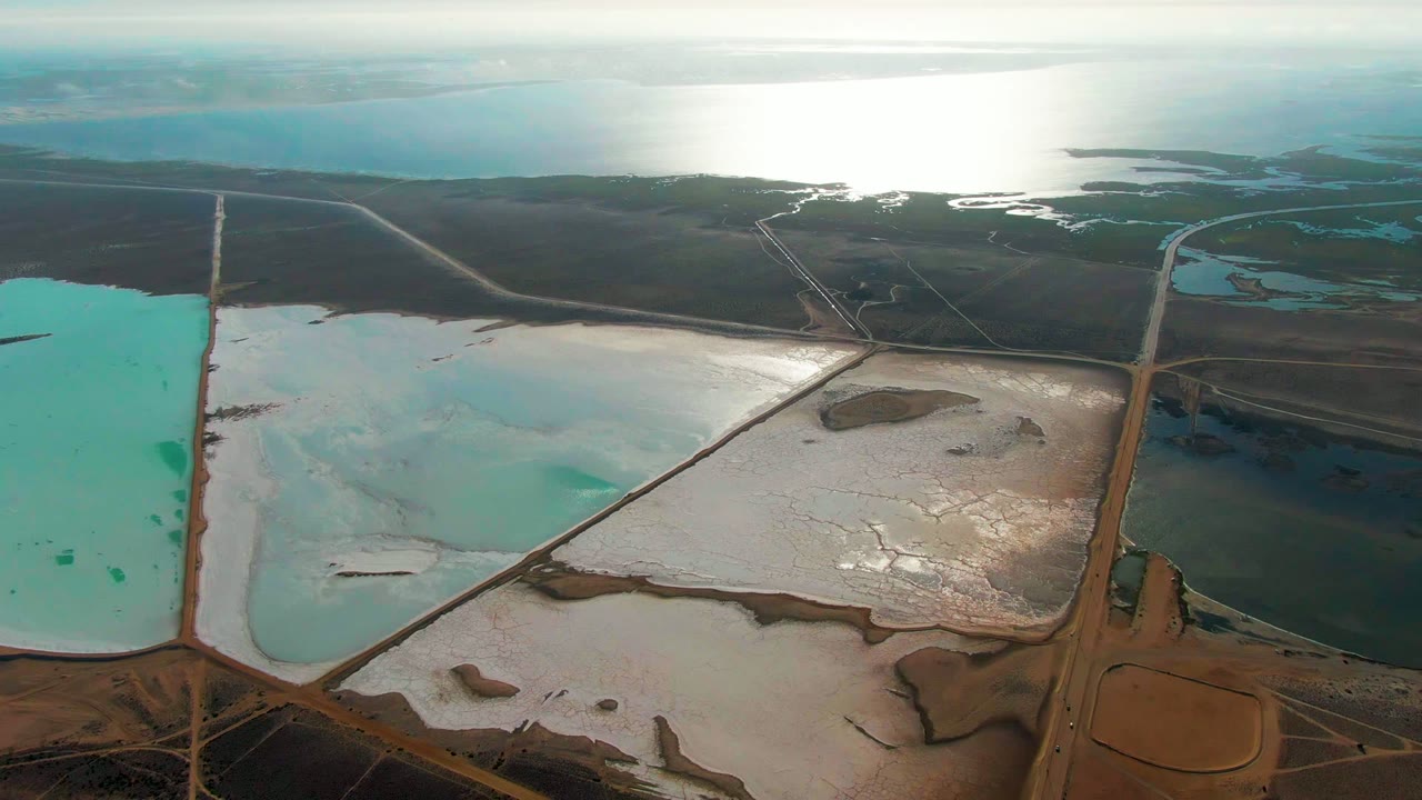 Aerial view in Guerrero Negro with a vertical turquoise evaporation pond at left, crusted white salt flats across the middle, road intersections below, dark terrain above, and no visible people.