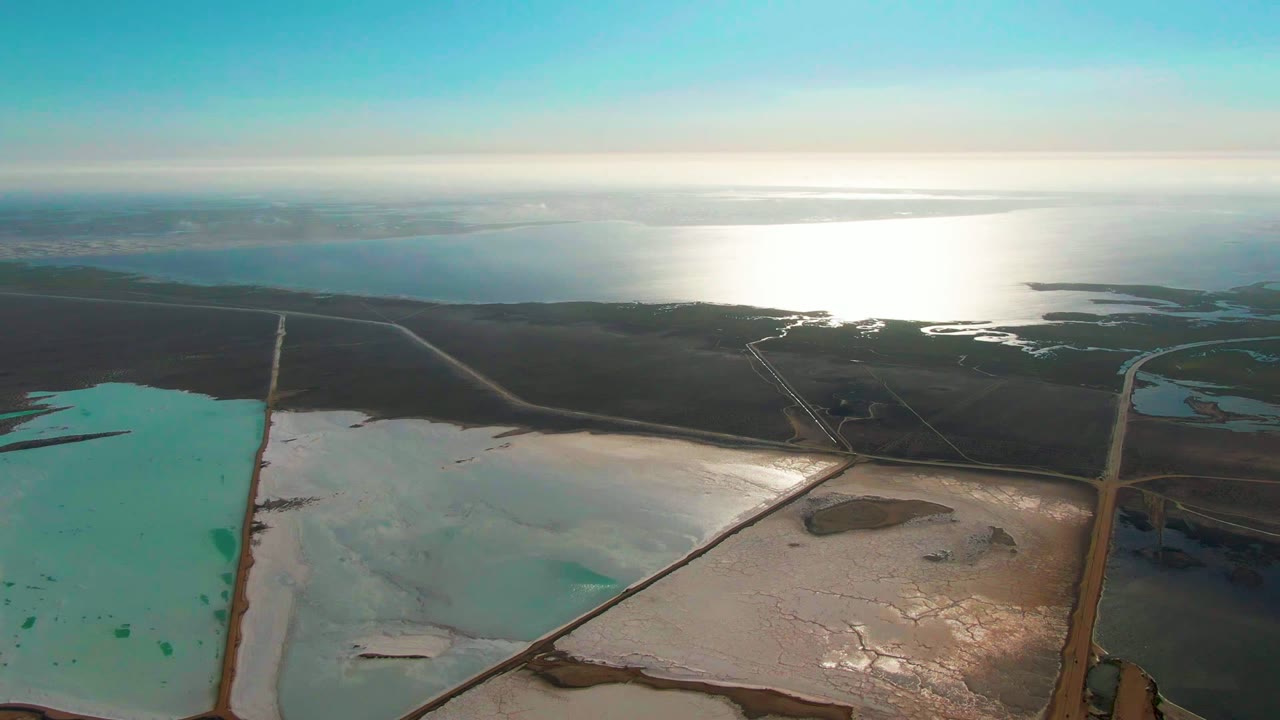 Opening aerial frame over Guerrero Negro salt evaporation ponds, with turquoise water at left, pale white basins at right, ocean on the horizon, bright sun reflection, and no visible people.