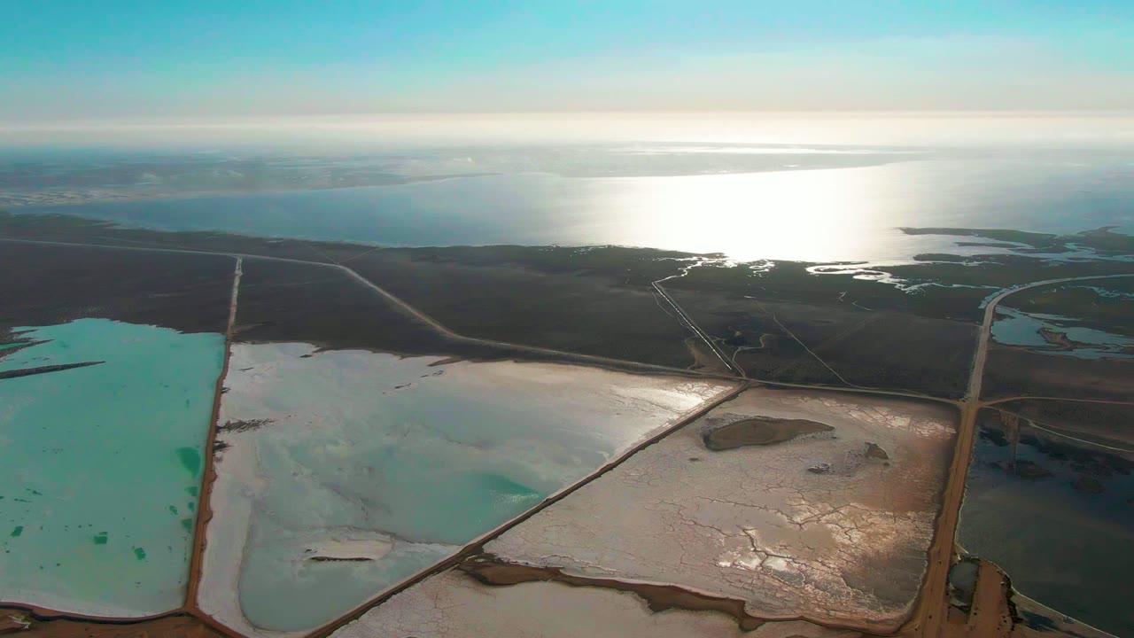 Drone view over rectangular salt ponds with bright ocean glare beyond dark land.