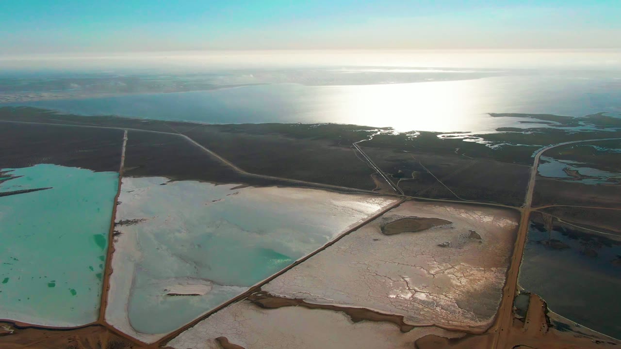 Mid-clip aerial frame over Guerrero Negro evaporation ponds, with a vertical turquoise basin, adjacent white pond, brown levee roads, distant ocean, and no visible people.