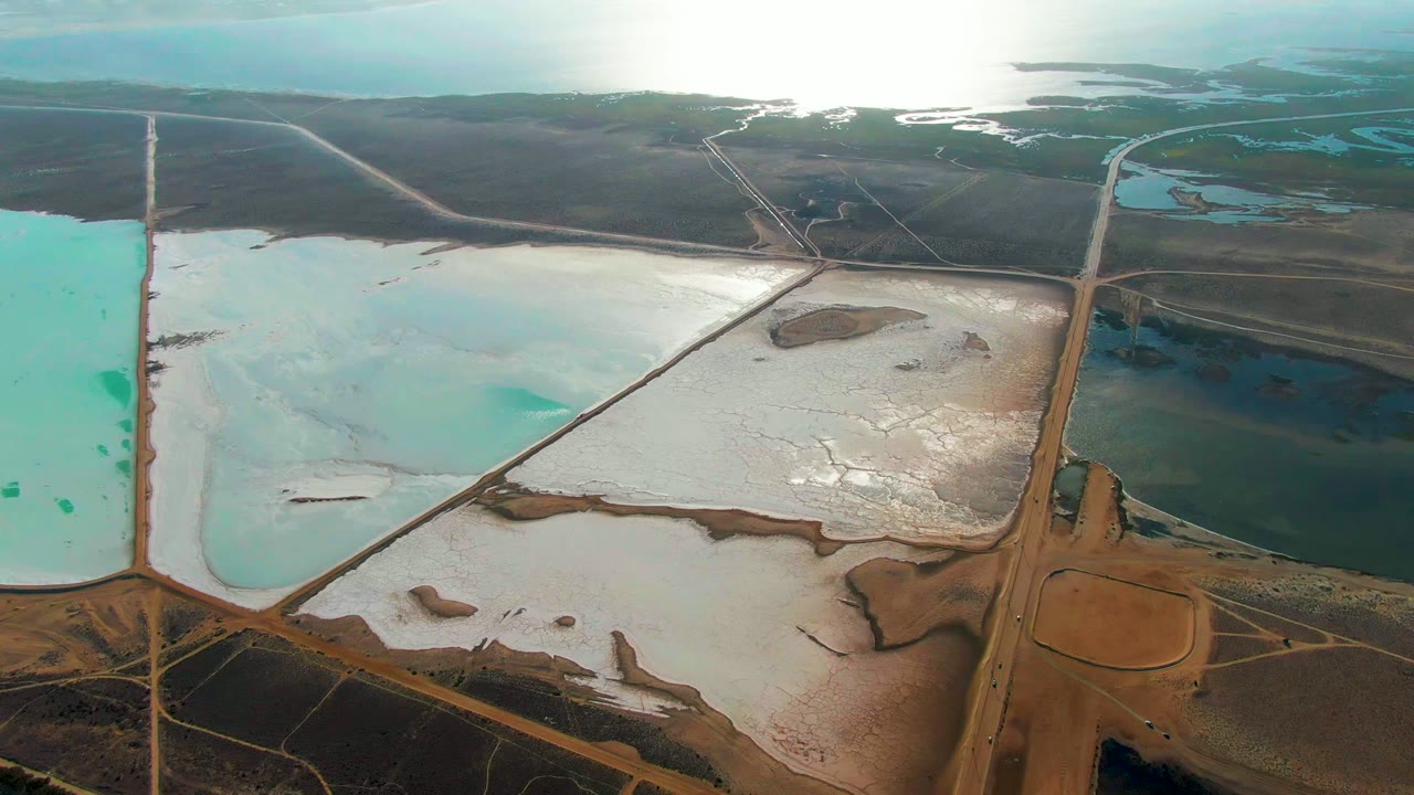 High aerial view over industrial salt evaporation ponds in Guerrero Negro, Baja California Sur, with turquoise water, white salt deposits, brown dikes, distant wetlands, and no visible people.