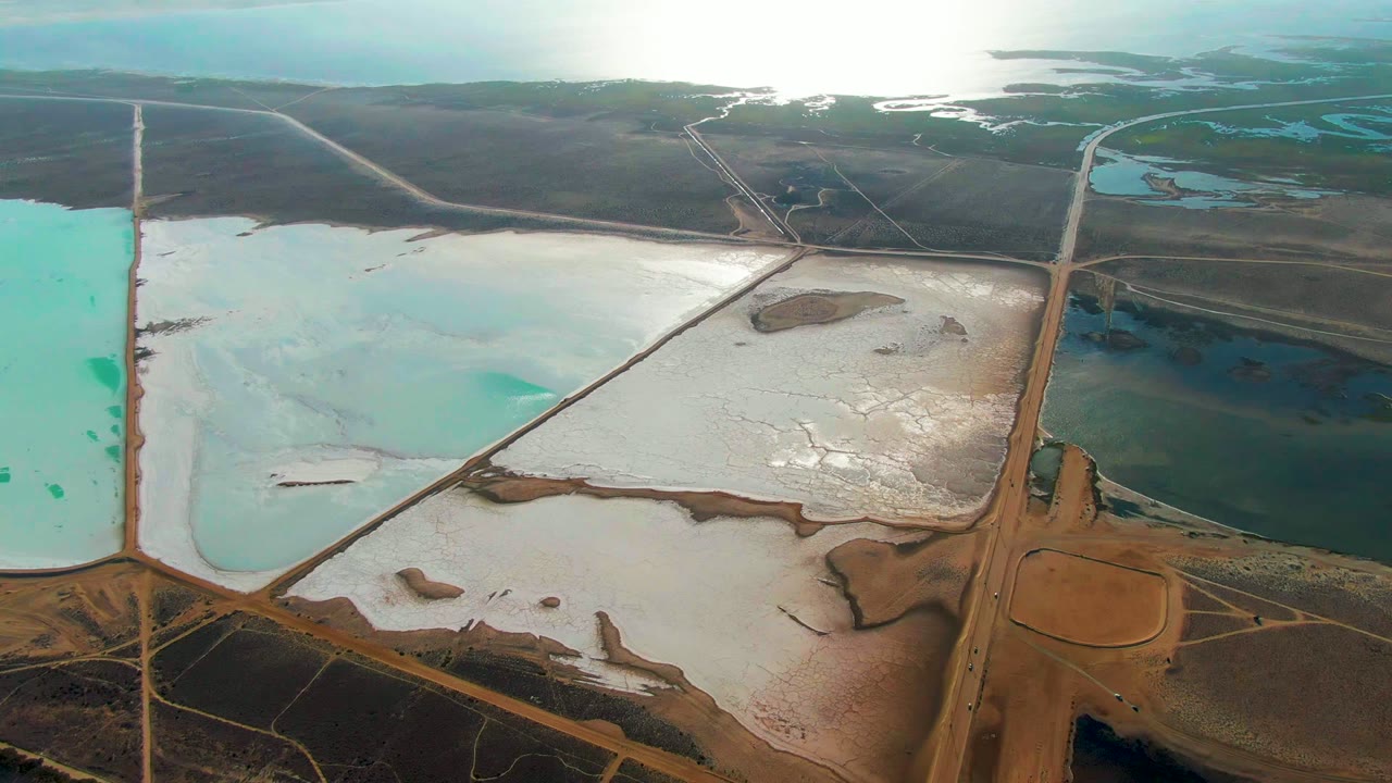 Aerial frame in Guerrero Negro showing a diagonal service road between pale white salt crust, shallow blue-green pond water, brown exposed earth, and no visible people.