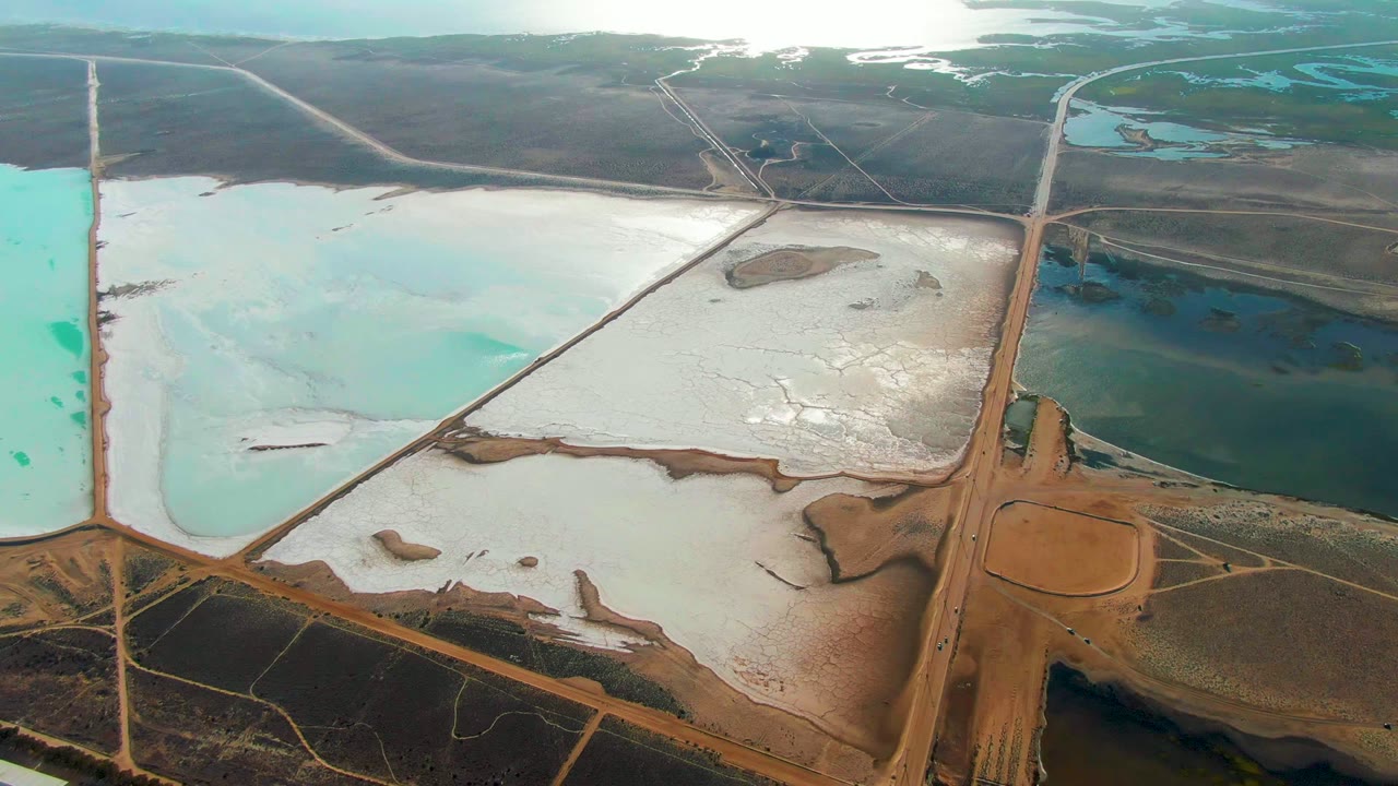 High aerial frame over salt evaporation ponds in Guerrero Negro, Mexico, with turquoise, white, brown, and dark green basins divided by dikes and no visible people.