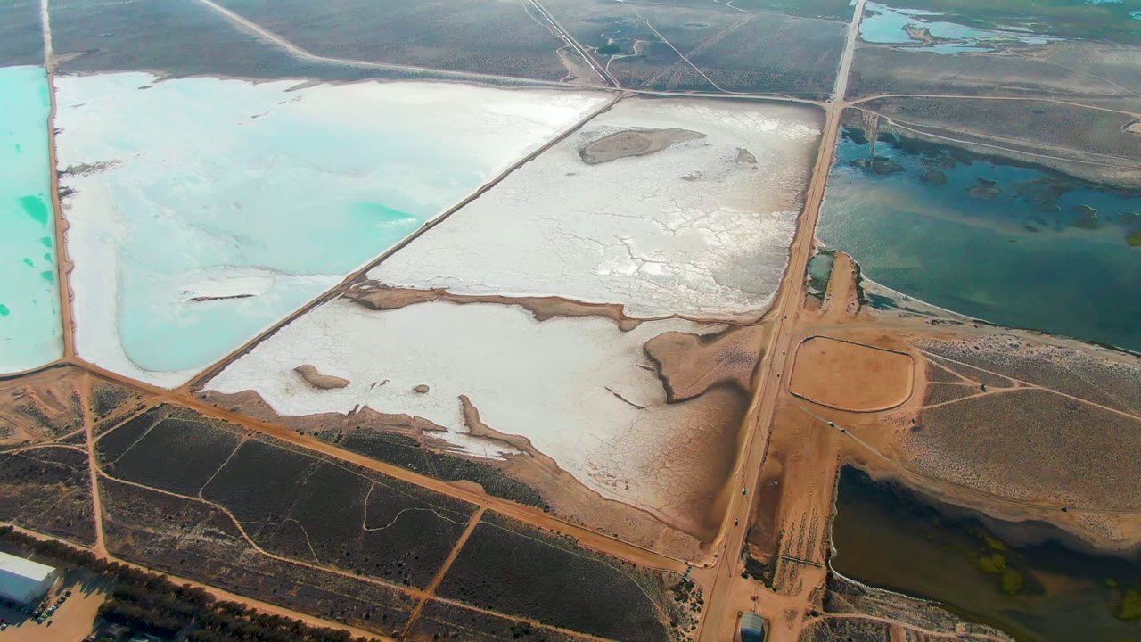 Opening aerial frame over Guerrero Negro salt evaporation ponds, with white flats, turquoise water basins, a central dirt road, a tiny truck shape, and no visible people.