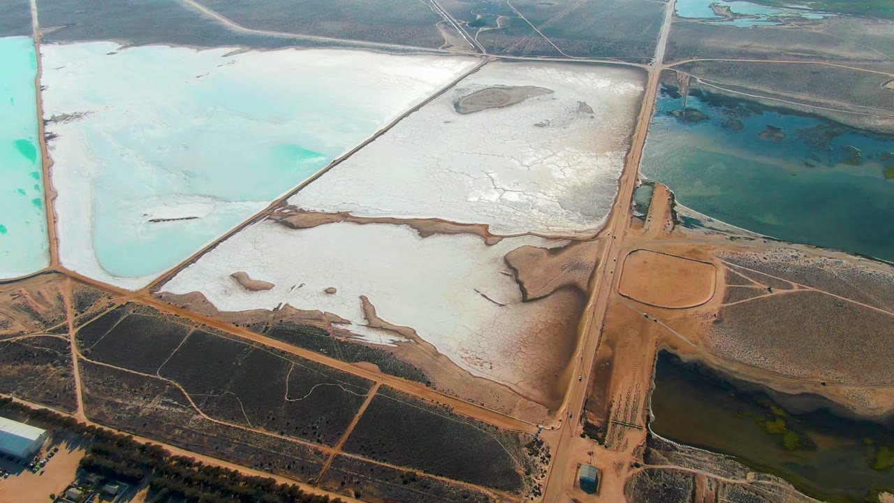 At two seconds, the Guerrero Negro aerial view holds on white salt flats and turquoise ponds divided by a tan road, with the small truck still on the road and no visible people.