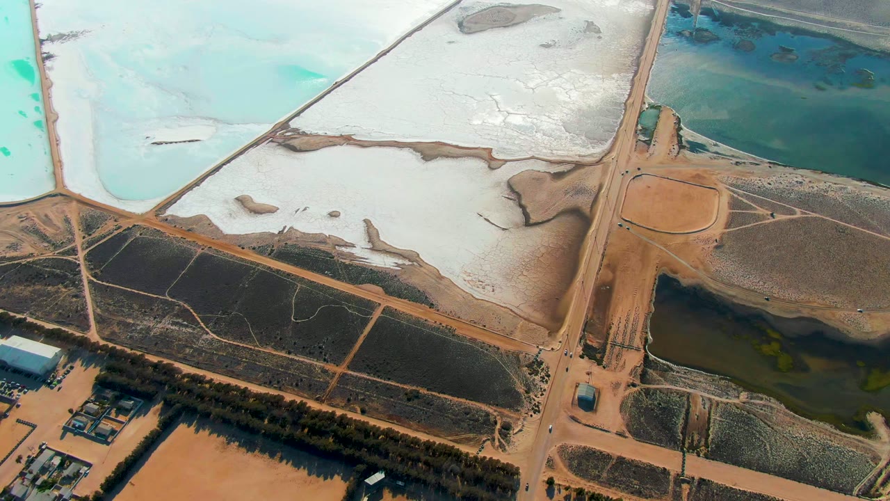 Overhead view in Guerrero Negro, Baja California Sur, showing pale salt ponds, turquoise water, dark brine areas, dirt roads, and no visible people at the start of the clip.