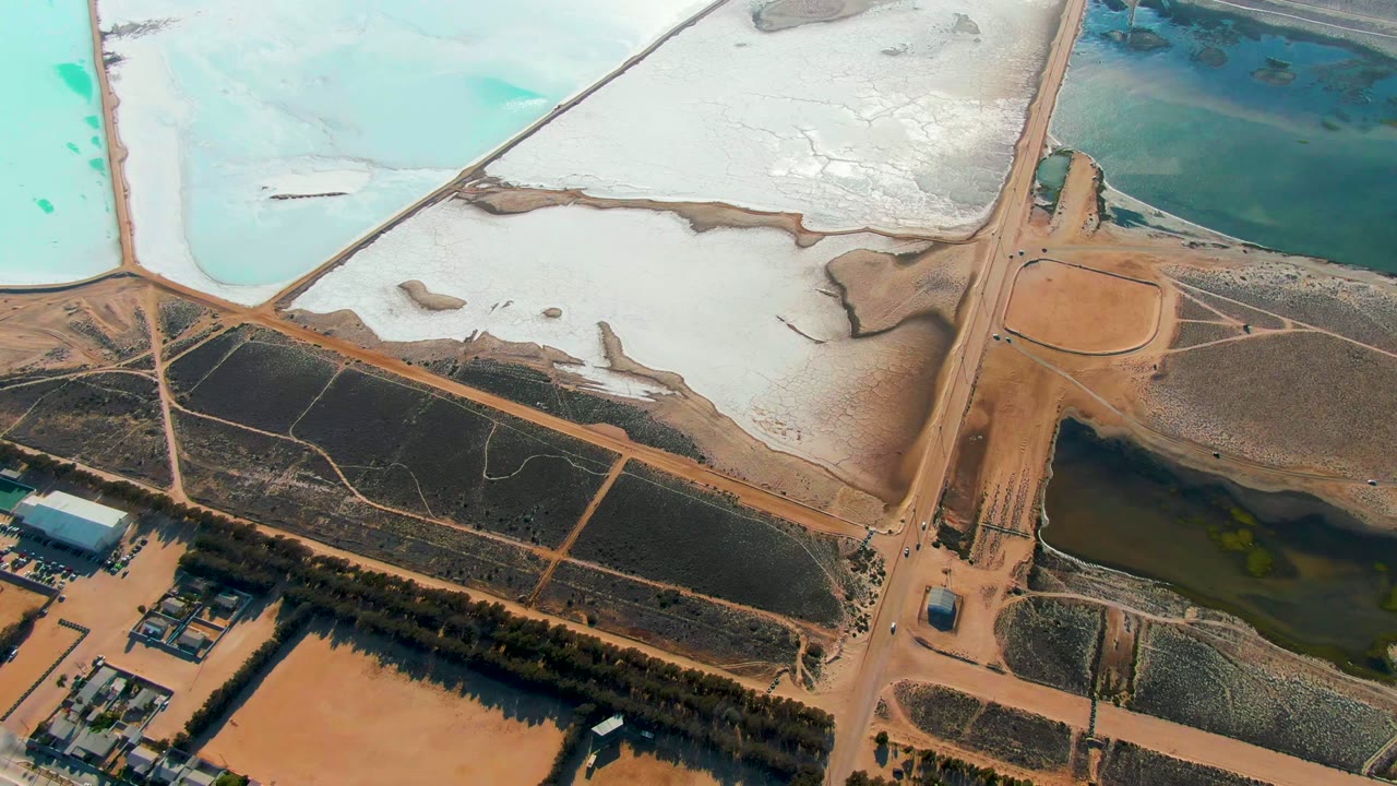 High drone view in Guerrero Negro with bright turquoise ponds at the top, white mineral flats, brown roads, a dark pond near the lower edge, and no visible people.