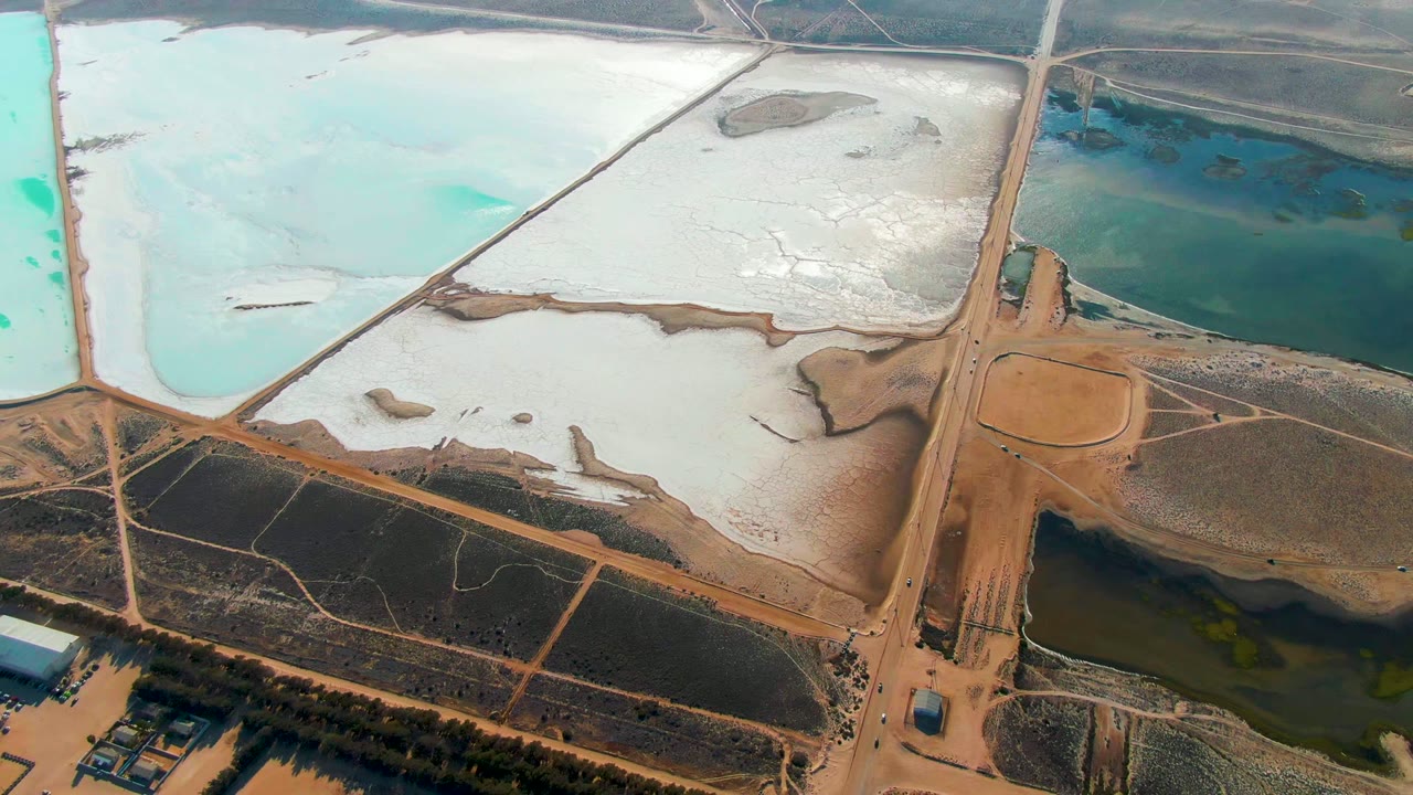 Opening frame of Guerrero Negro salt evaporation ponds with turquoise, white, dark blue, and brown basins divided by tan roads, no visible people.