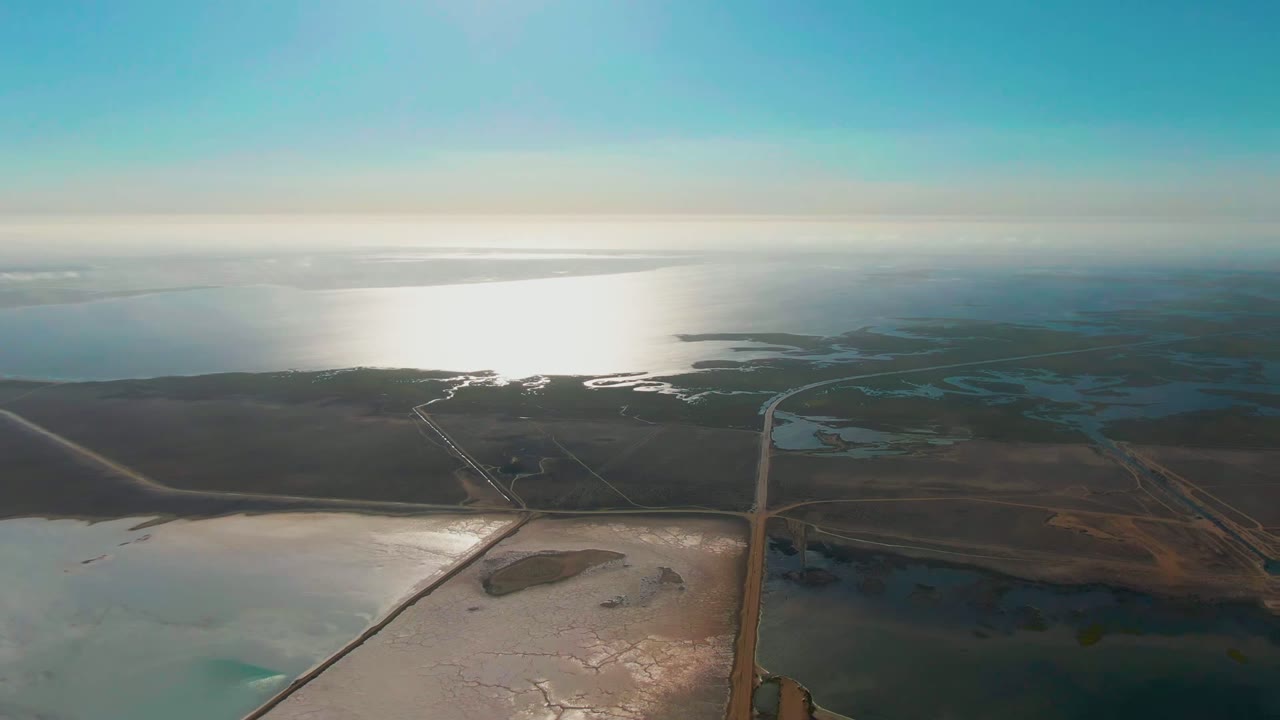 Aerial view over Guerrero Negro salt evaporation ponds and coastal water, with a bright sun reflection on the ocean, straight road lines below, blue sky above, and no visible people.