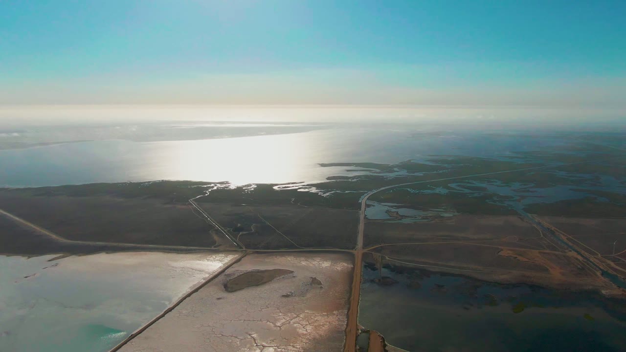 Aerial view of Guerrero Negro coastal evaporation ponds, with the ocean reflecting hard light near the horizon, low wetlands and roads in the middle distance, and no visible people.