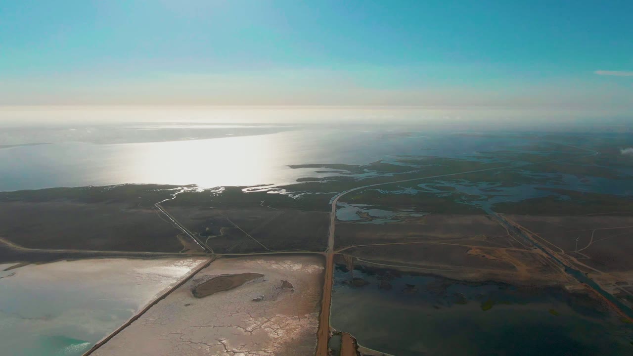 Opening aerial frame over Guerrero Negro, Baja California Sur, with bright sea reflection on the left, shallow salt-flat water below, brown wetlands, a straight road through the center, and no visible people.
