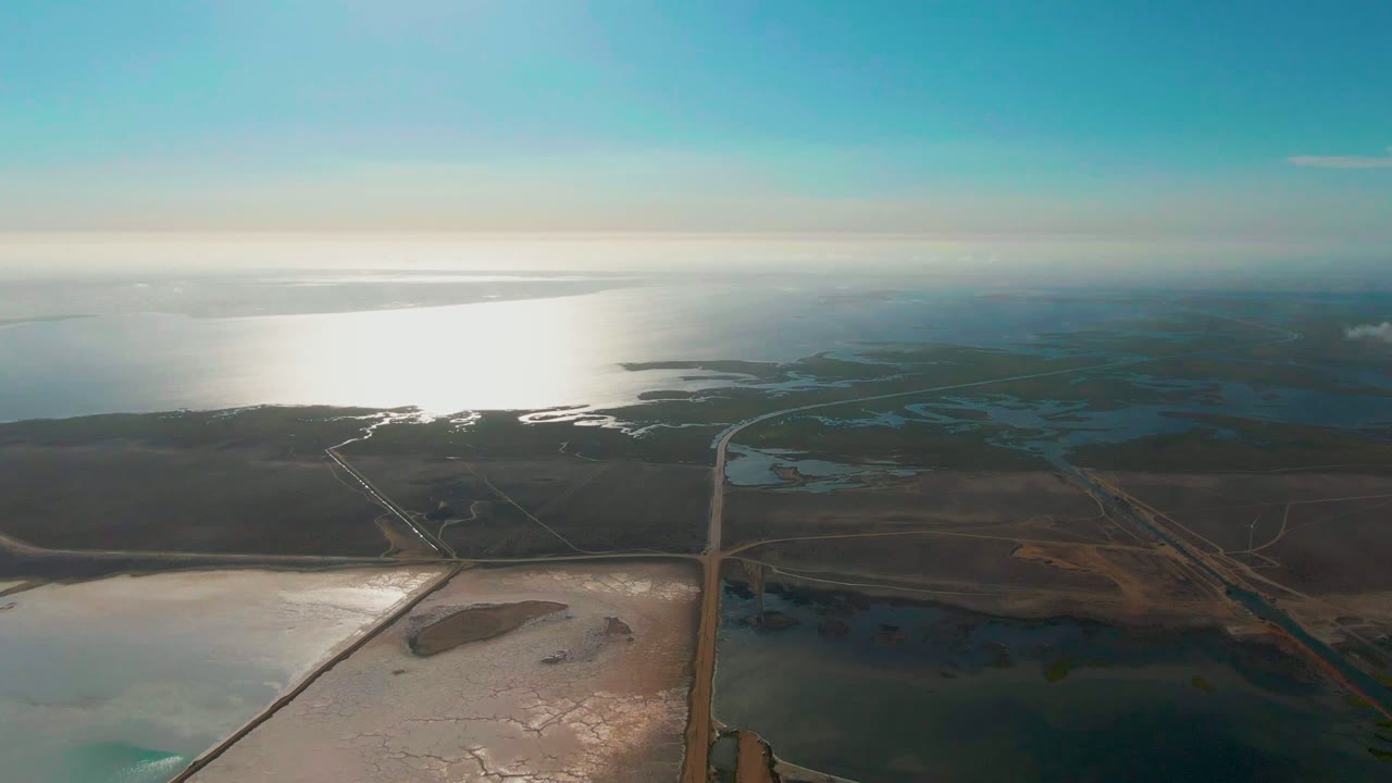 High coastal view with sun glare on water, wetlands, salt-flat pools, and a road.