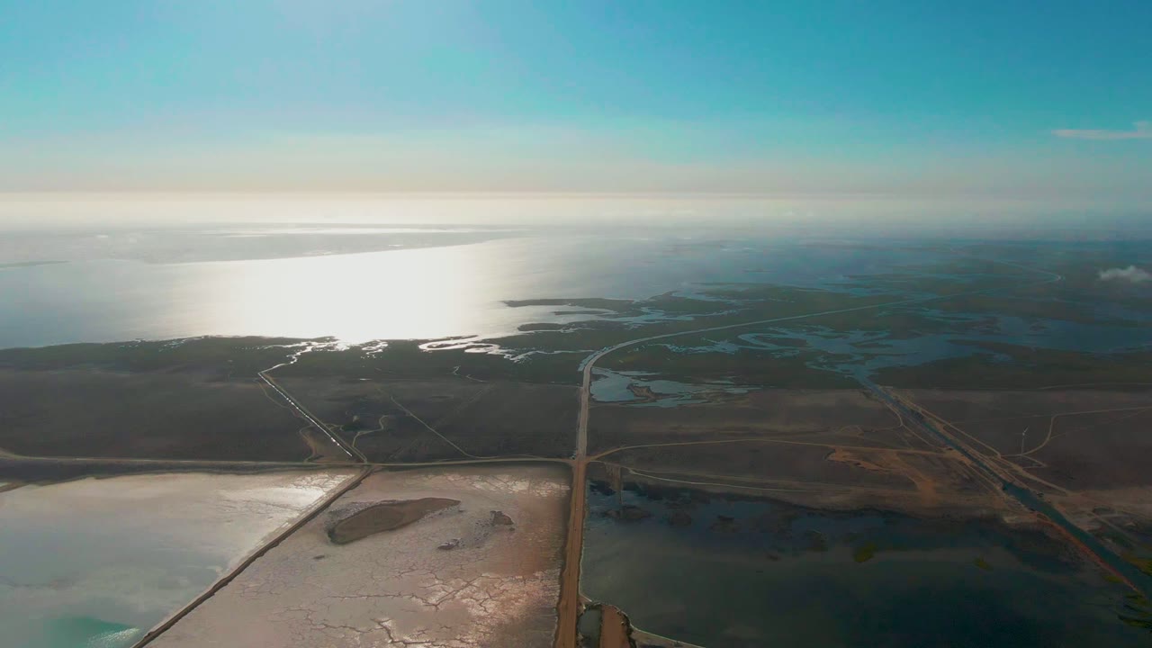 At 0:02, the aerial composition shows the road running upward through brown wetland terrain toward reflective coastal water, with pale salt-flat water in the foreground and no visible people.