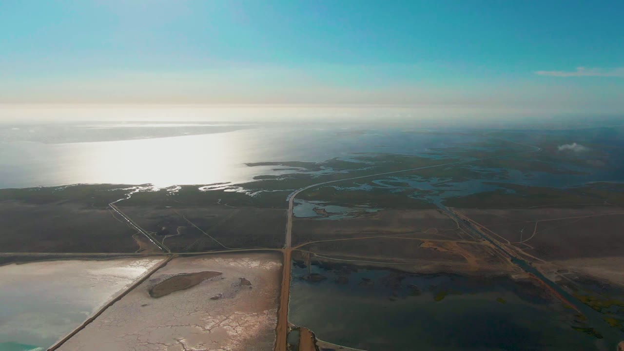Aerial view over Guerrero Negro, Baja California Sur, with bright ocean water on the left, a straight road crossing salt ponds below, dry brown land beyond, and no visible people.