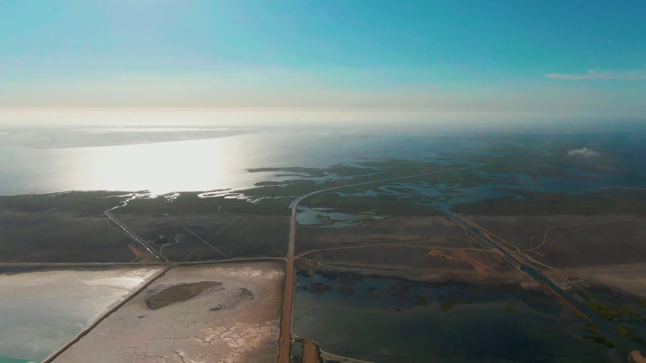 High aerial view of salt flats, ocean glare, road, and distant coastline.