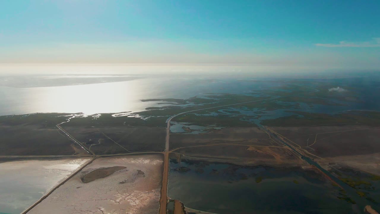 Wide aerial view of Guerrero Negro coastal salt flats with blue-green pond areas in the midground, brown dry land, bright ocean water to the left, and no visible people.