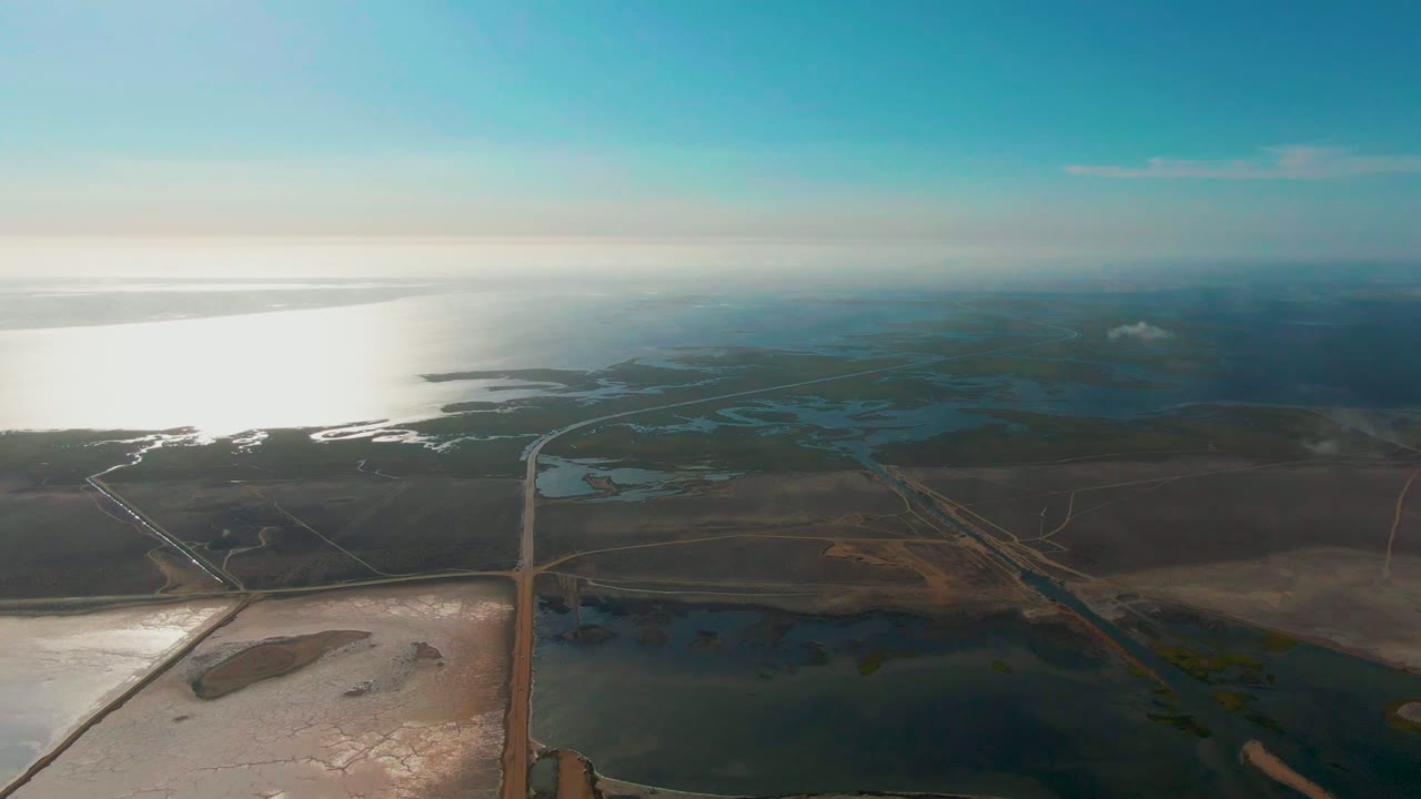 At 0:00, a high aerial view over Guerrero Negro shows a straight road running through coastal wetlands, reflective water, open fields, and no visible people.