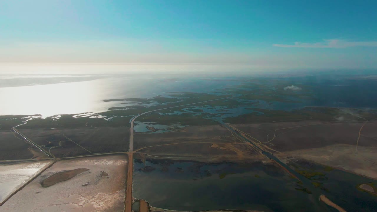 At 0:02, the aerial view shows the vehicle continuing along the road through Guerrero Negro wetlands, with water channels, open land, and no visible people.