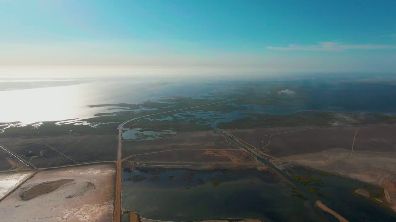 High aerial view over Guerrero Negro coastal wetlands with dark water in the foreground, flat brown land, thin roads, bright sea reflection at left, blue sky, and no visible people.