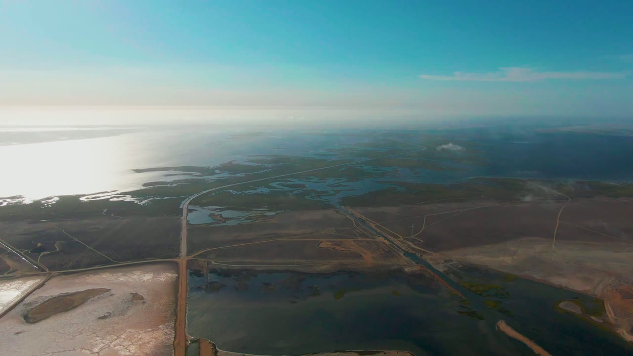 Wide aerial wetlands with water channels, road lines, and distant sea glare.
