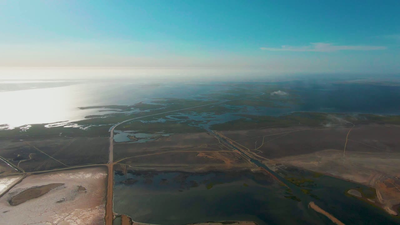 Wide drone view over Guerrero Negro wetlands with a pale salt flat at lower left, dark lagoon water below, road lines through brown land, distant sea, hazy horizon, and no visible people.