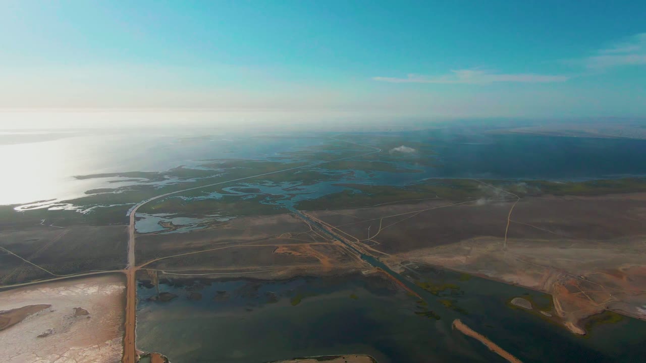 Opening aerial frame over Guerrero Negro, Baja California Sur, with a straight road entering from the lower left, shallow water and dry flats below, hazy horizon, and no visible people.
