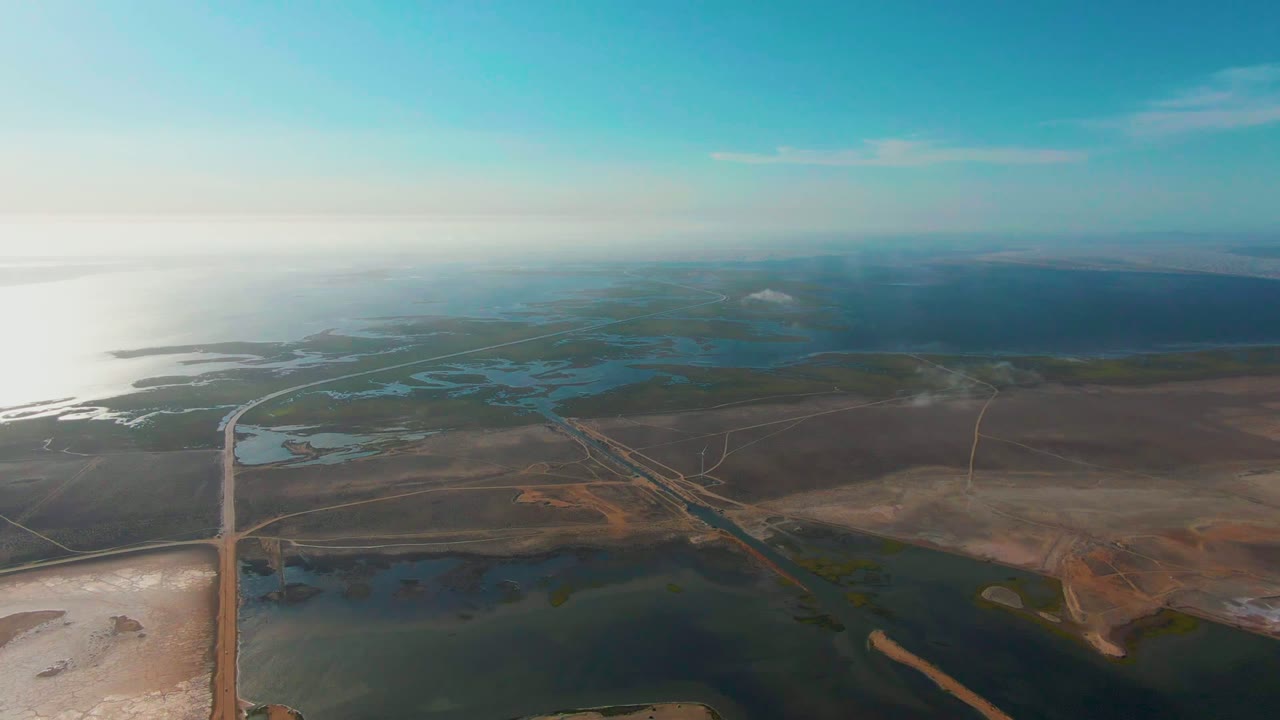 Mid-clip aerial over Guerrero Negro, Baja California Sur, showing the long road crossing wetland pools, dry earth patches, distant water, and no visible people.