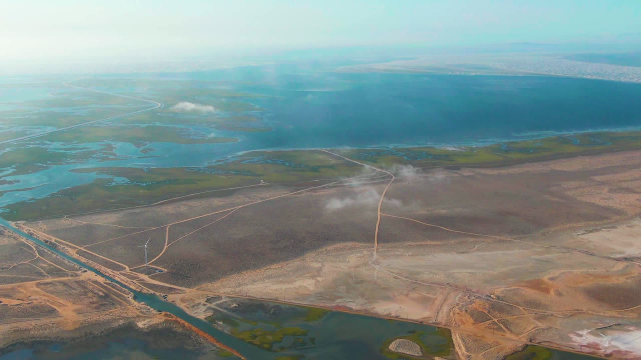 Opening aerial frame over Guerrero Negro, Baja California Sur, Mexico, with blue water beyond dry brown terrain, pale roads, wetland channels, a white mist plume near center, and no visible people.